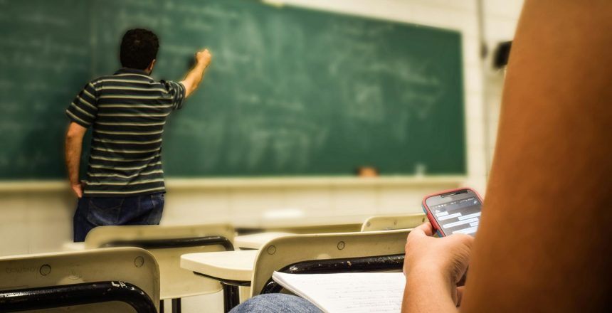 FEatured image showing a student in a classroom on her phone while the teacher writes on a chalkboard. Many students appear absent.